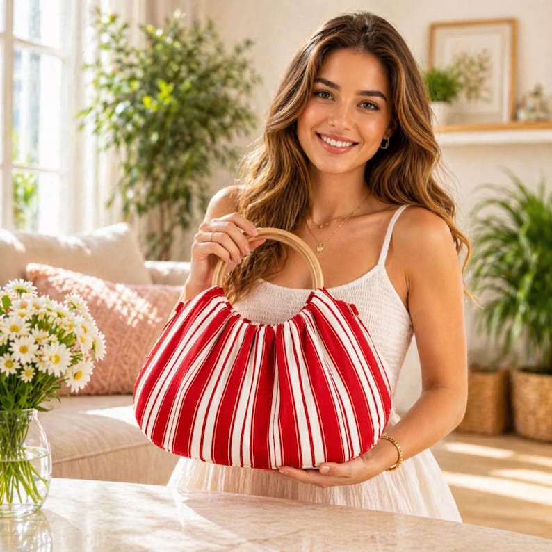 May include: A red and white striped handbag with a wooden handle. The bag is held by a person wearing a white dress. The bag's design features vertical red stripes separated by white stripes. The handle is a light brown color.