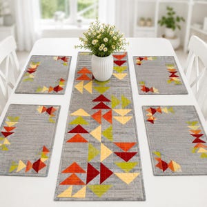 May include: A quilted table runner and four matching rectangular placemats in grey fabric, each featuring a geometric pattern of small triangles in shades of red, orange, yellow, and lime green. A white vase with white flowers sits on top.