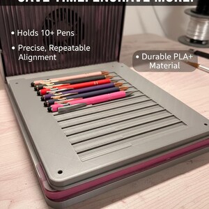 May include: A gray pen holder with a pink trim, holding multiple pens of various colors. The text on the image reads "SAVE TIME. ENGRAVE MORE." and highlights features like "Holds 10+ Pens" and "Durable PLA+ Material."