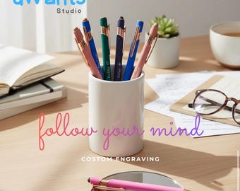 May include: A collection of colorful pens in a white ceramic holder, with the text "follow your mind" and "custom engraving". A pink pen rests on a small mirror. The pens are blue, green, and pink.