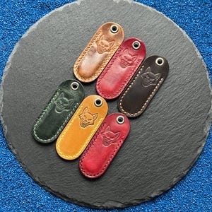 Könnte beinhalten: Sechs Leder-Schlüsselanhänger in verschiedenen Farben, darunter Braun, Rot und Grün. Jeder Schlüsselanhänger hat ein Fuchsmotiv und eine Metallöse zum Befestigen von Schlüsseln. Sie sind auf einer dunkelgrauen, kreisförmigen Oberfläche angeordnet.