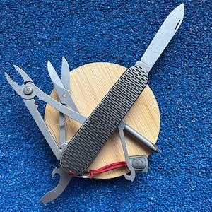 May include: A multi-tool pocket knife with a textured dark gray handle and a silver blade extended. The tool features integrated pliers, scissors, a bottle opener, and a screwdriver, with a small red cord attached to the base of the handle.