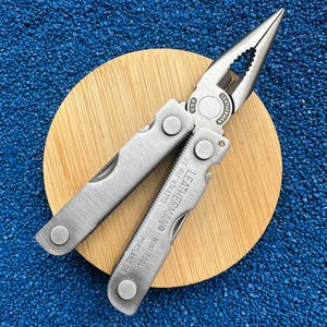 Könnte beinhalten: Ein silberfarbenes Leatherman Mini Tool Multifunktionswerkzeug mit Spitzzange und klappbaren Griffen. Der Metallkörper ist mit dem Text LEATHERMAN MINI TOOL PORTLAND OR und US PAT 4744272 graviert, mit USA-Prägung nahe dem Zangengelenk.
