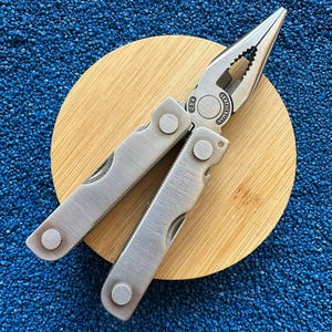 May include: A silver Leatherman Mini Tool multi-tool with needle-nose pliers, featuring the text Leatherman Mini Tool and US Pat 4227261 engraved on the handle, with a brushed metal finish and visible pivot screws and joints.
