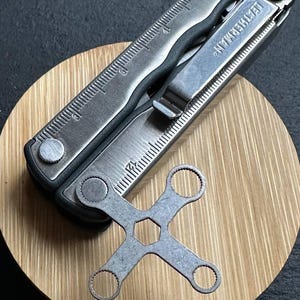 Op de afbeelding: Een zilverkleurig multitool met een liniaal en een zakclip, plus een aparte moersleutel. De multitool is gemarkeerd met "LEATHERMAN". De gereedschappen worden gepresenteerd op een ronde, lichtbruine houten ondergrond.