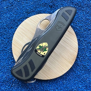 以下が含まれることがあります： シルバーの刃が付いた、濃い緑色のスイスアーミーナイフ。ハンドルには黒いラバーグリップとビクトリノックスのロゴが付いています。ナイフには「Sanität」の文字が入った小さなエンブレムが付いています。