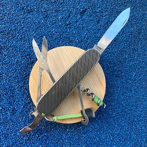 Puede incluir: Una navaja multiusos con mango texturizado gris oscuro y hoja plateada desplegada. La herramienta incluye tijeras plateadas, un abrebotellas y un sacacorchos con un cordón verde brillante sujeto a una pequeña anilla metálica en la base del mango.