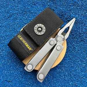 Könnte beinhalten: Ein silbernes Leatherman-Multitool mit Spitzzange und einer schwarzen Nylontasche. Die Tasche verfügt über einen Metalldruckknopf mit eingeprägtem Leatherman-Logo und den in gelbem Garn gestickten Markennamen auf der vorderen Klappe.