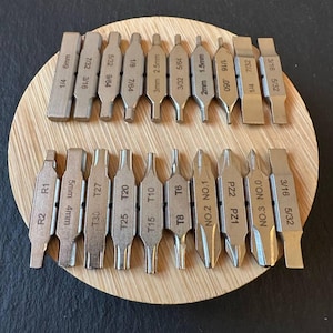 以下が含まれることがあります： 丸い木製のベースに配置された、銀色の金属製ドライバービットのセット。 ビットには、1/4、3/16、6mm、2mmなどのサイズを含む、インチとミリメートルでの測定値が記載されています。 さまざまな種類のネジに対応しています。