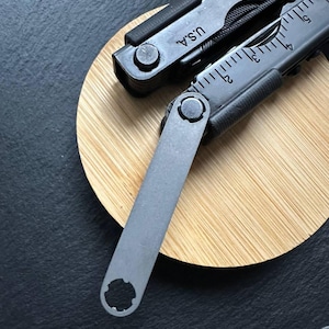 Könnte beinhalten: Ein schwarzes Multitool mit dem Text "U.S.A." und einem Lineal auf der Seite. Das Werkzeug ist teilweise geöffnet und zeigt ein graues, flaches Werkzeug mit einer kreisförmigen Öffnung. Das Werkzeug liegt auf einer hellbraunen, kreisförmigen Holzoberfläche.
