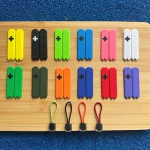 Puede incluir: Una colección de tiradores de cremallera y repuestos coloridos en varios colores: amarillo, negro, verde, azul, rosa, blanco, naranja, rojo y morado. Cada tirador tiene un signo más y se muestra sobre una superficie de madera. Medidas en cm.