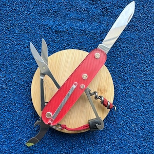 Puede incluir: Una navaja multiusos con mango rojo translúcido y componentes de metal plateado, que incluye una hoja principal, tijeras, un sacacorchos y un abrebotellas. La herramienta está colocada sobre una base circular de madera.