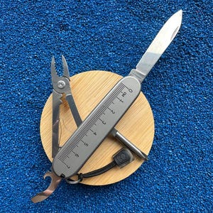 May include: A silver multi-tool pocket knife with a 7-centimetre ruler printed on the handle. The tool features an extended blade, pliers, a bottle opener, and a screwdriver. A black lanyard with a small logo tag is attached to the base of the handle.