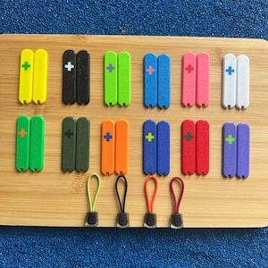 Puede incluir: Una colección de coloridos tiradores de cremallera en varios tonos, incluyendo amarillo, negro, verde, azul, rosa, naranja, rojo y morado. Cada tirador presenta un signo más y está emparejado con un cordón a juego y un conector negro.
