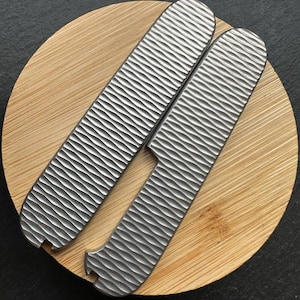 Pode incluir: Duas pegas de faca de metal prateado texturizado com um padrão de ondas, repousando sobre uma tábua de corte redonda de madeira castanha clara. As pegas têm uma forma curva com um entalhe numa extremidade. O fundo é escuro.