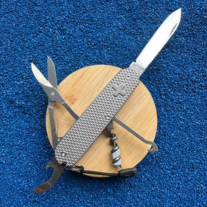 Könnte beinhalten: Ein silbernes Multifunktions-Taschenmesser mit strukturiertem Metallgriff und einem Kreuz-Emblem. Das Werkzeug ist geöffnet und zeigt eine Klinge, eine Schere, einen Korkenzieher und einen Flaschenöffner auf einer runden, hellen Holzscheibe.