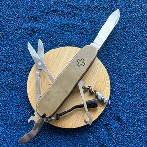 Puede incluir: Una navaja multiusos con mango de bronce metálico y el emblema de la cruz suiza. La herramienta incluye una hoja desplegada con la marca Victorinox, tijeras abiertas, un abrebotellas, un sacacorchos y un cordón negro.