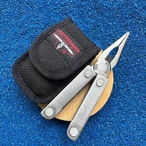 May include: A silver stainless steel Leatherman multi-tool with needle-nose pliers and a black nylon carrying case. The tool features the Leatherman logo and USA patent text engraved on the handle, while the case displays a red and white logo.