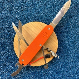 Puede incluir: Una navaja multiusos con cachas texturizadas de color naranja brillante y un emblema de cruz suiza. La navaja cuenta con una hoja de acero inoxidable desplegada, tijeras abiertas, un sacacorchos y un abrebotellas. Incluye un pequeño cordón naranja.