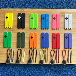 Puede incluir: Una colección de tiradores de cremallera coloridos y pequeñas herramientas, dispuestas sobre una superficie de madera. Los tiradores de cremallera vienen en varios colores, incluyendo amarillo, negro, verde, azul, rosa, blanco, verde, naranja, rojo y morado, cada uno con un signo más.