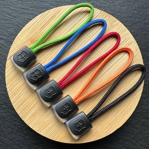 Può includere: Cinque tiretti per cerniera colorati in verde, blu, rosso, arancione e nero. Ogni tiretto ha una base in plastica nera con un logo e un anello di corda dello stesso colore. Sono disposti su una superficie rotonda di legno.