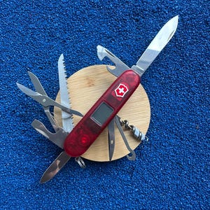 May include: A red translucent multi-tool pocket knife featuring a digital display, a white cross emblem, and multiple extended stainless steel implements including a large blade, scissors, a saw, a corkscrew, and various screwdrivers.
