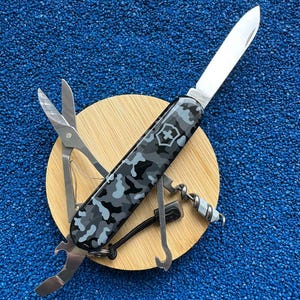 Puede incluir: Una navaja multiusos con un mango de patrón de camuflaje en gris, negro y blanco. La navaja se muestra con la hoja principal extendida, junto con unas tijeras, un abrebotellas, un sacacorchos y una pequeña herramienta de gancho visible.