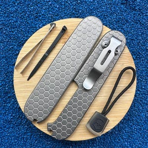 Puede incluir: Un juego de multiherramientas que incluye dos cachas de metal gris con un patrón hexagonal de panal, un clip de bolsillo plateado unido a una cacha, unas pinzas plateadas, un palillo de plástico negro y un cordón negro con un dije.