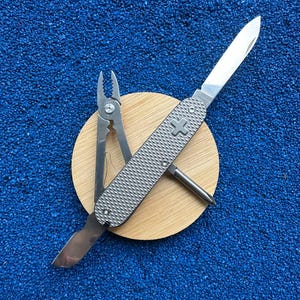 May include: A silver multi-tool pocket knife with a textured metallic handle featuring a cross emblem. The tool is open, displaying a sharp blade, pliers, a screwdriver, and a small flat tool, all resting on a circular light wood base.