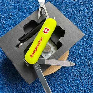 Op de afbeelding: Een gele en zilveren multitool met de tekst "Emergency Tool" en een rood kruislogo. De tool bevat een schaar, een mes en een nagelvijl. Het wordt gepresenteerd in een zwarte doos op een blauw, getextureerd oppervlak.