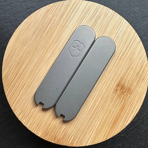 Puede incluir: Dos herramientas metálicas grises con una muesca en un extremo, apoyadas sobre una superficie redonda de bambú. Una herramienta tiene un logotipo. Las herramientas son probablemente para un propósito específico, posiblemente relacionado con una multiherramienta o cuchillo.