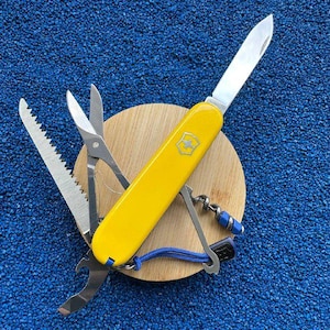 Puede incluir: Una navaja multiusos amarilla con hoja de acero inoxidable, tijeras, sierra para madera, abrebotellas y sacacorchos. La herramienta incluye un cordón azul y está colocada sobre una base circular de madera de color claro.