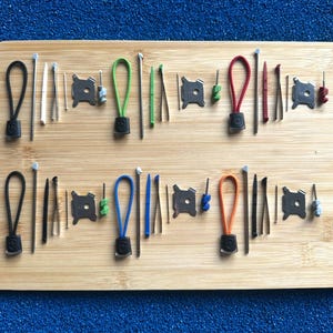 Könnte beinhalten: Eine Sammlung von Ersatzteilen für Multitools auf einem Holzbrett, bestehend aus verschiedenfarbigen Kordelzügen, Metallstiften, Pinzetten, kleinen Kunststoffwerkzeugen und quadratischen Metallkomponenten mit mittigen Löchern.