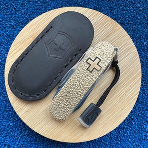 Op de afbeelding: Een goud-zilveren Zwitsers zakmes met een getextureerde handgreep en een zwart koord. Het mes heeft een wit kruis embleem en wordt geleverd met een zwarte leren schede. Het mes ligt op een houten oppervlak.