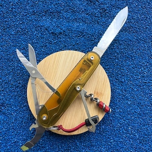 Puede incluir: Una navaja multiusos con mango de color ámbar translúcido que incluye una hoja dentada, tijeras, un sacacorchos con detalles rojos, un abrebotellas y un destornillador pequeño, colocada sobre una base circular de madera clara.