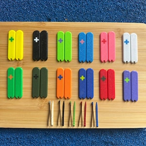 Puede incluir: Una colección de objetos de plástico rectangulares y coloridos, cada uno con un signo más, dispuestos sobre una superficie de madera. Los objetos son de varios colores, incluyendo amarillo, negro, verde, azul, rosa, blanco, naranja, rojo y morado.
