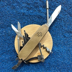 Puede incluir: Una navaja multiusos con mango de metal en tono bronce y un emblema de cruz. La herramienta está parcialmente desplegada, mostrando una hoja plateada, tijeras, un sacacorchos, un abrebotellas y un accesorio de destornillador extendido.