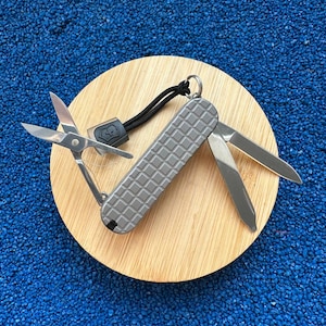 Puede incluir: Una navaja multiusos gris con un patrón de rejilla texturizado en el mango. La herramienta cuenta con unas tijeras de metal plateado abiertas y una hoja de metal plateado desplegada. Incluye un cordón negro con un pequeño dije cuadrado.