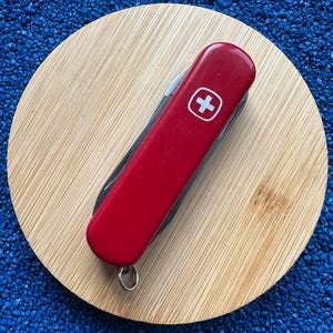 May include: A red Swiss Army knife with a white cross emblem, resting on a round, light brown wooden coaster. The knife is closed, showing its red handle and a small metal loop. The background is a textured blue surface.