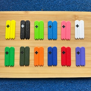 Puede incluir: Una colección de pequeños objetos rectangulares de colores, cada uno con un signo más. Los objetos son de varios colores, incluyendo amarillo, negro, verde, azul, rosa, blanco, verde oliva, naranja, rojo y morado. Están dispuestos sobre una superficie de madera.