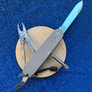 Puede incluir: Una multiherramienta suiza Victorinox de color beige con mango texturizado y emblema de cruz suiza. La herramienta cuenta con una hoja dentada desplegada, alicates, destornillador y abrebotellas, todo fabricado en acero inoxidable pulido.