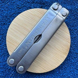 Könnte beinhalten: Ein silbernes Leatherman Multi-Tool mit dem Markennamen auf der Seite eingraviert. Das Werkzeug liegt auf einem runden, hellen Holzuntersetzer vor einem blauen, strukturierten Hintergrund. Das Werkzeug ist geschlossen und hat eine Vielzahl von Funktionen.
