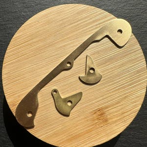 Könnte beinhalten: Nahaufnahme von vier messingfarbenen Metall-Uhrwerksteilen. Die Teile umfassen ein langes, dünnes Stück mit gebogenem Ende und zwei Löchern sowie drei kleinere Teile mit kreisförmigen Löchern. Sie liegen auf einer runden, hellen Holzoberfläche.
