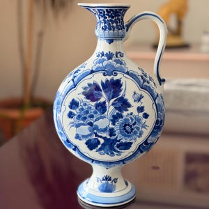May include: A white and blue ceramic pitcher with a floral design. The pitcher has a round body, a handle, and a spout. The floral design is in shades of blue and white. The pitcher is on a pedestal base.