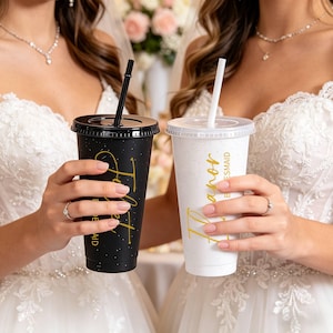 May include: Two glitter-covered tumblers, one black and one white, with gold script. The black tumbler reads "To Be A Maid" and the white tumbler reads "Honor Be A Maid". Both have black straws and lids. Wedding theme.