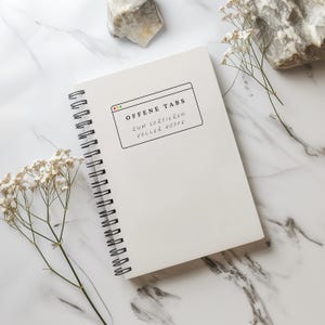 Könnte beinhalten: Eine weiße Keramik-Tasse mit schwarzem Henkel und dem Text "But First, Coffee" in fetter, schwarzer Schrift. Die Tasse steht vor einem verschwommenen Hintergrund, der eine gemütliche Umgebung suggeriert.