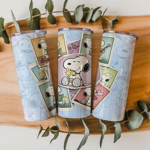 May include: Three stainless steel tumblers featuring Snoopy illustrations. The tumblers have a light blue background with Snoopy character outlines. Each tumbler has a different Snoopy design, with images of Snoopy on cards.
