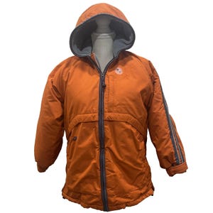 May include: An orange hooded jacket with a gray interior lining and a full-length zipper. The jacket has gray stripes on the sleeves and a small logo on the chest. The jacket is designed for cold weather.