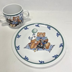 May include: A white ceramic plate and mug set featuring a teddy bear design. The plate has a green border with blue leaf accents and a teddy bear illustration. The mug has a matching design with a teddy bear and a blue and white checkered pattern.