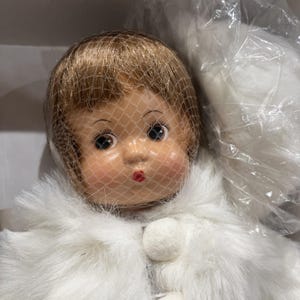 Puede incluir: Una muñeca vintage con la cara pintada, que lleva un abrigo de piel sintética blanco y un velo de red. La muñeca tiene el pelo castaño, mejillas rosadas y labios rojos. El abrigo se abrocha con un botón de pompón blanco.